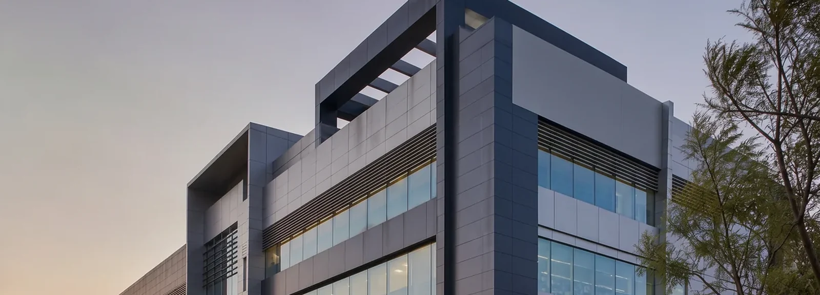 Modern commercial building clad in grey and navy panels with a long glazed frontage at dusk