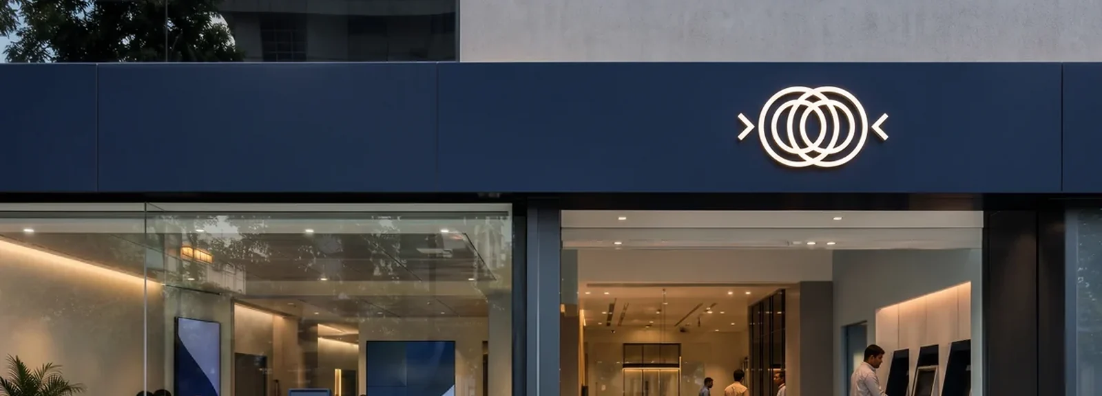 Bank branch frontage with a navy fascia band, an illuminated brand mark and ATMs visible through the glass