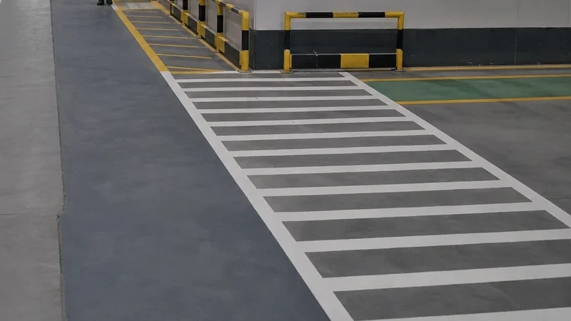White hatched pedestrian crossing and lane lines on a grey warehouse floor, yellow and black safety barriers behind